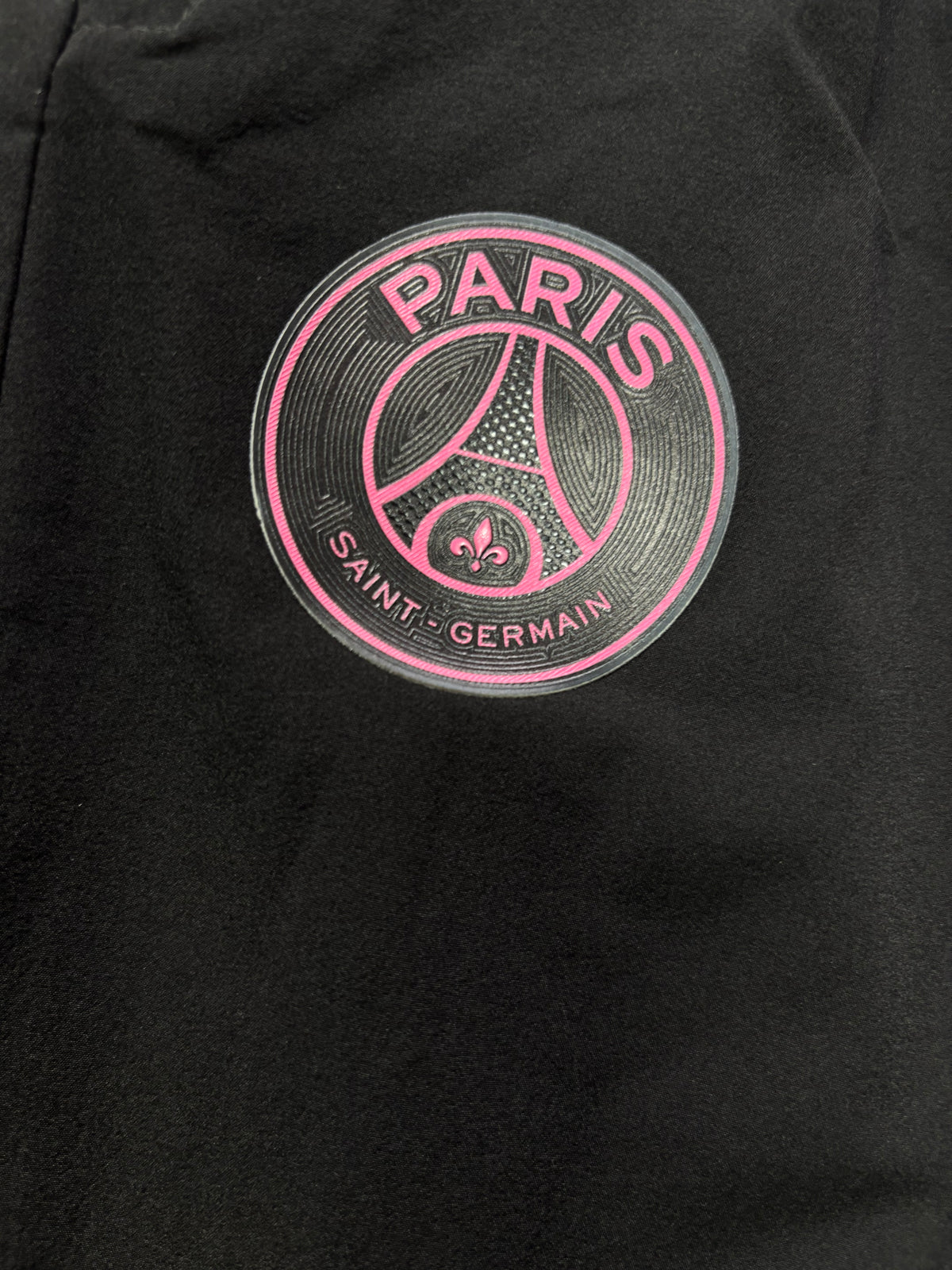 Close-up of a black garment featuring a circular Paris Saint-Germain logo printed in pink and gray on the chest.